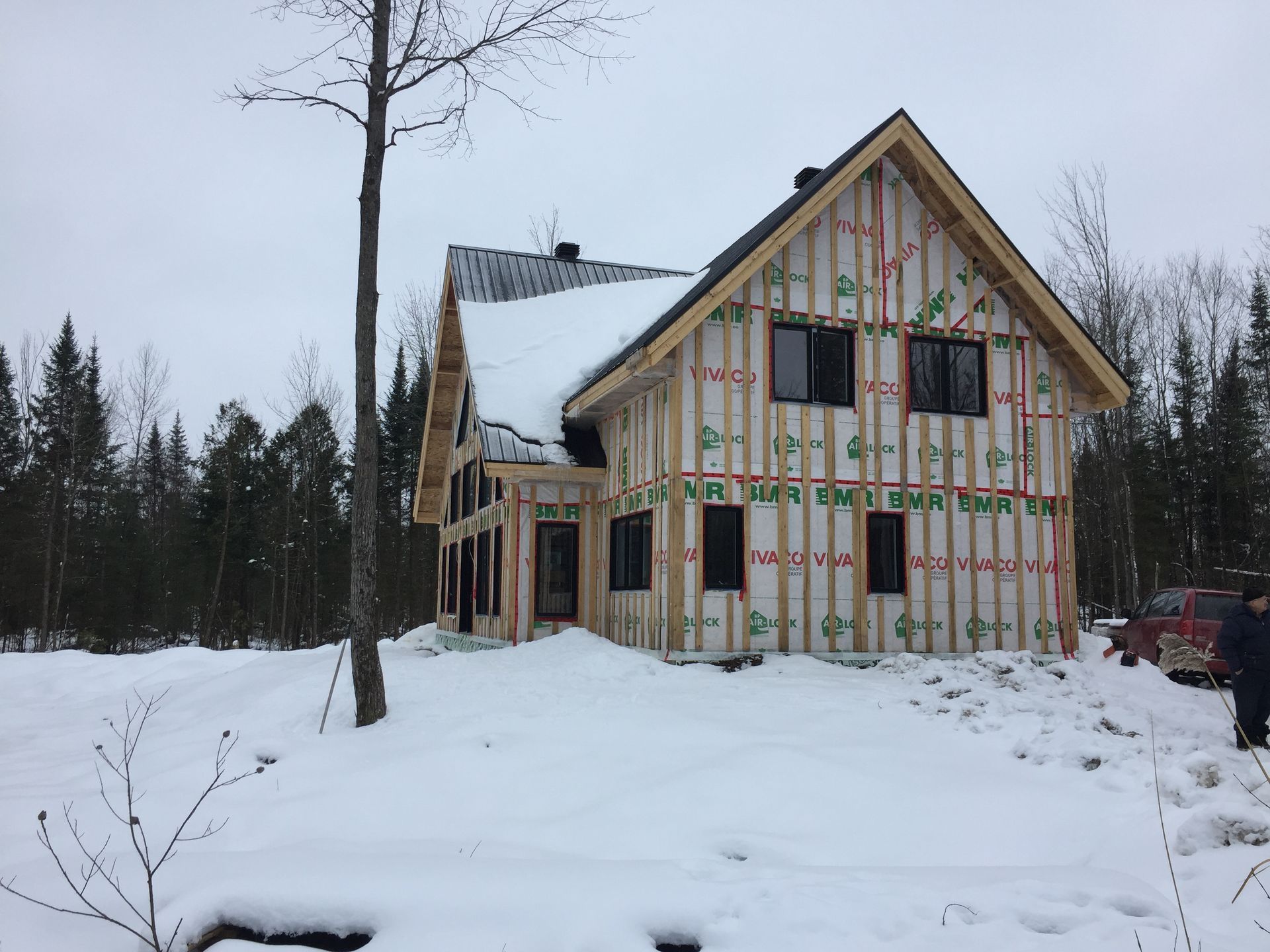 Maison en construction en hiver, entourée de neige et d'arbres. Charpente en bois apparente.
