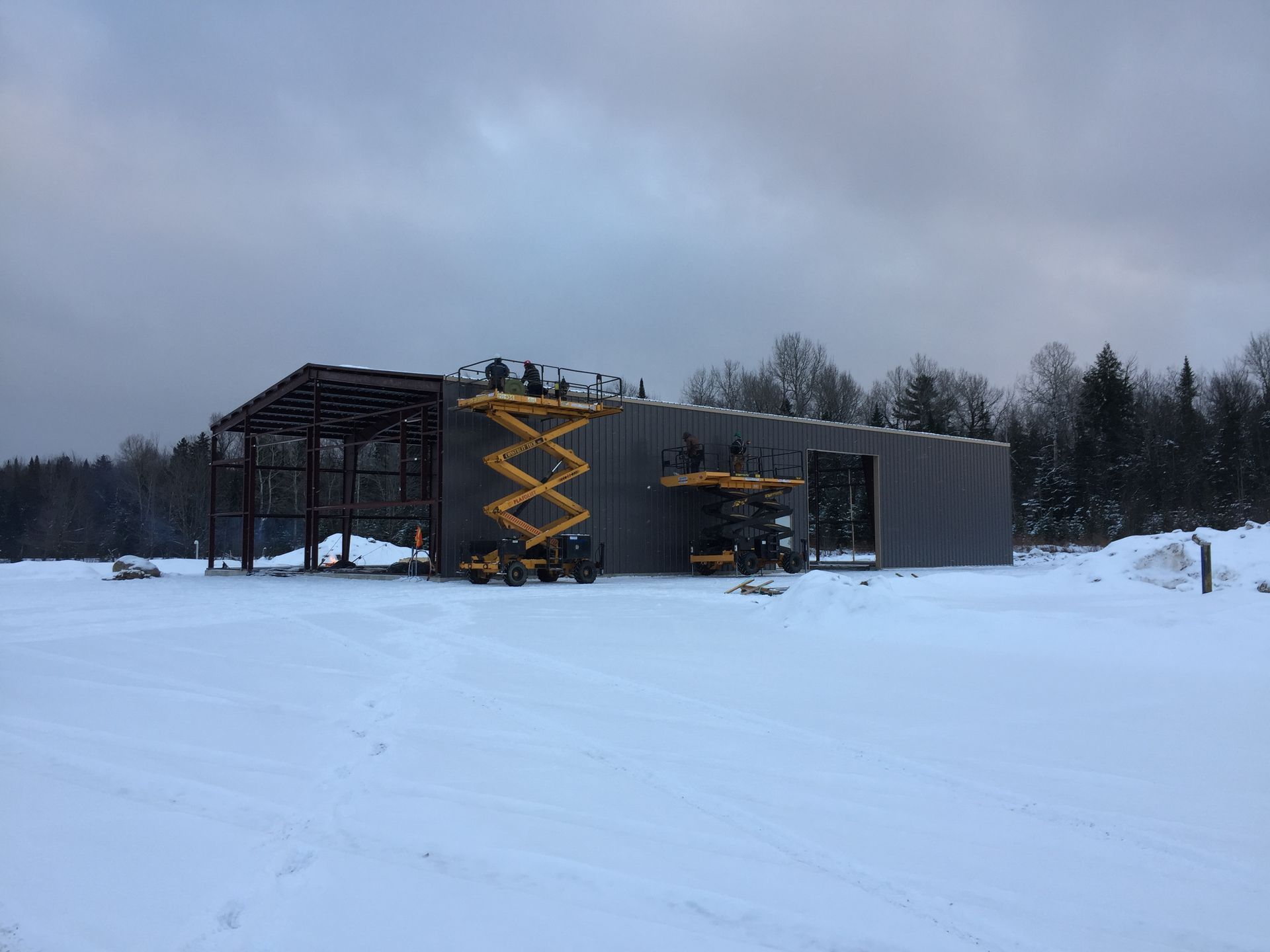 Construction d'un bâtiment métallique gris dans un champ enneigé ; ouvriers sur une nacelle élévatrice, ciel nuageux.