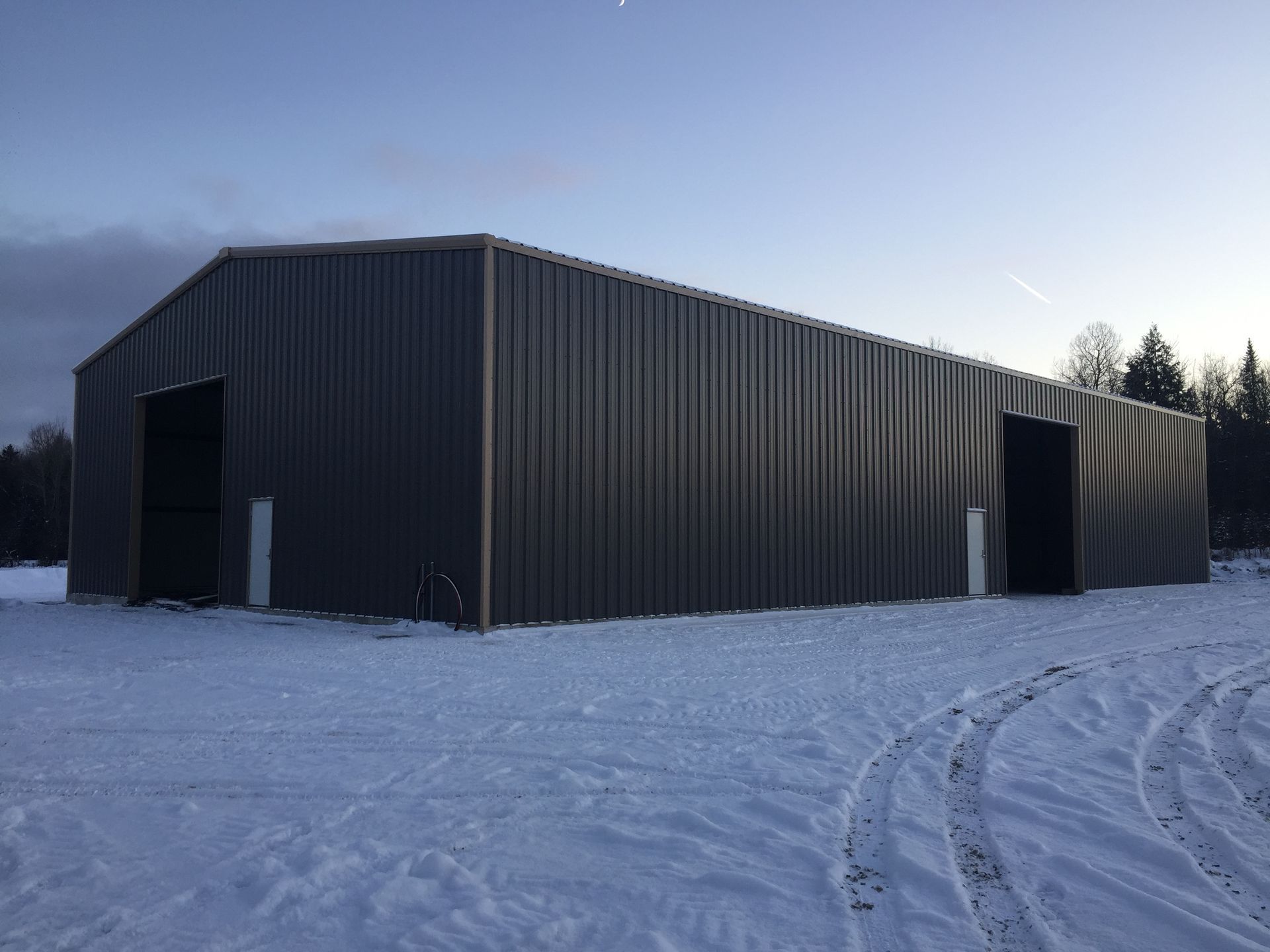 Grange en métal gris avec deux portes ouvertes, dans un champ enneigé au crépuscule.