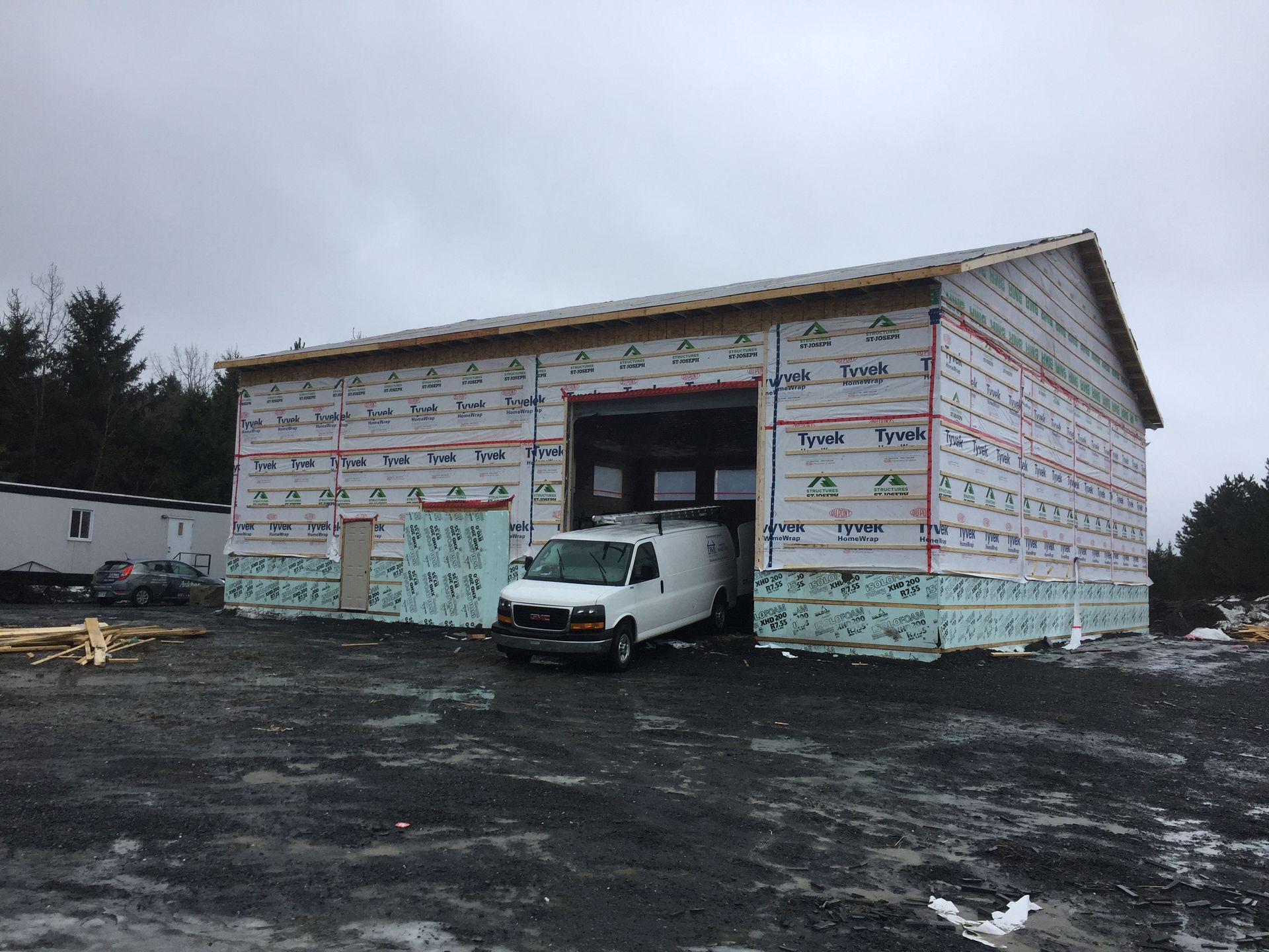 Une camionnette blanche pénètre dans un bâtiment en construction. La charpente est apparente et le bâtiment est recouvert d'un film Tyvek. Le ciel est gris et couvert.