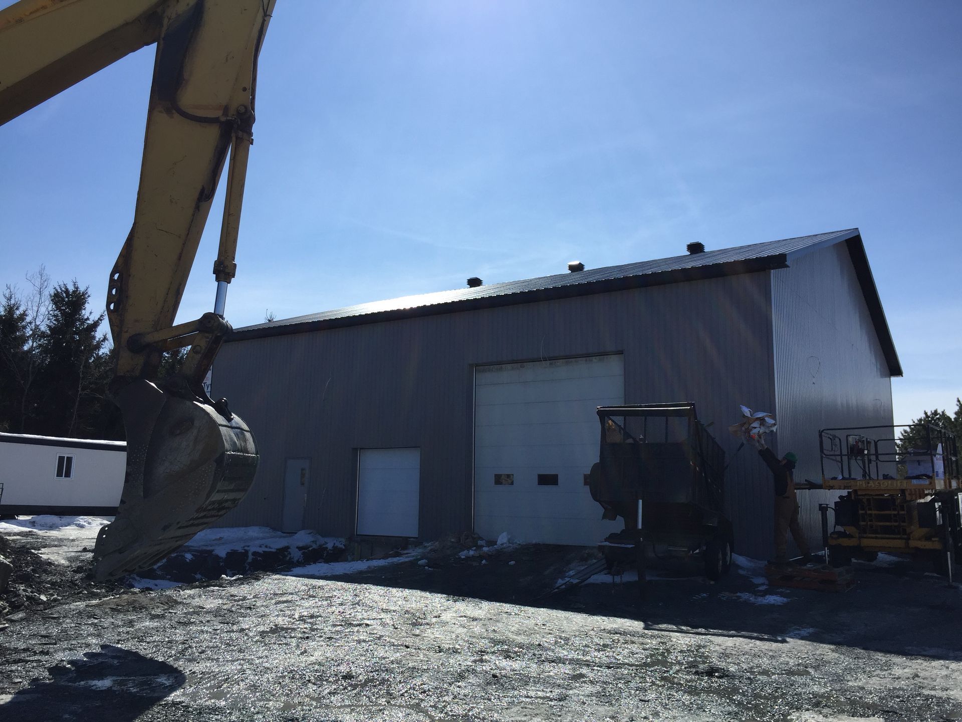 Un godet jaune d'excavatrice devant un bâtiment industriel métallique gris avec une porte de garage fermée.