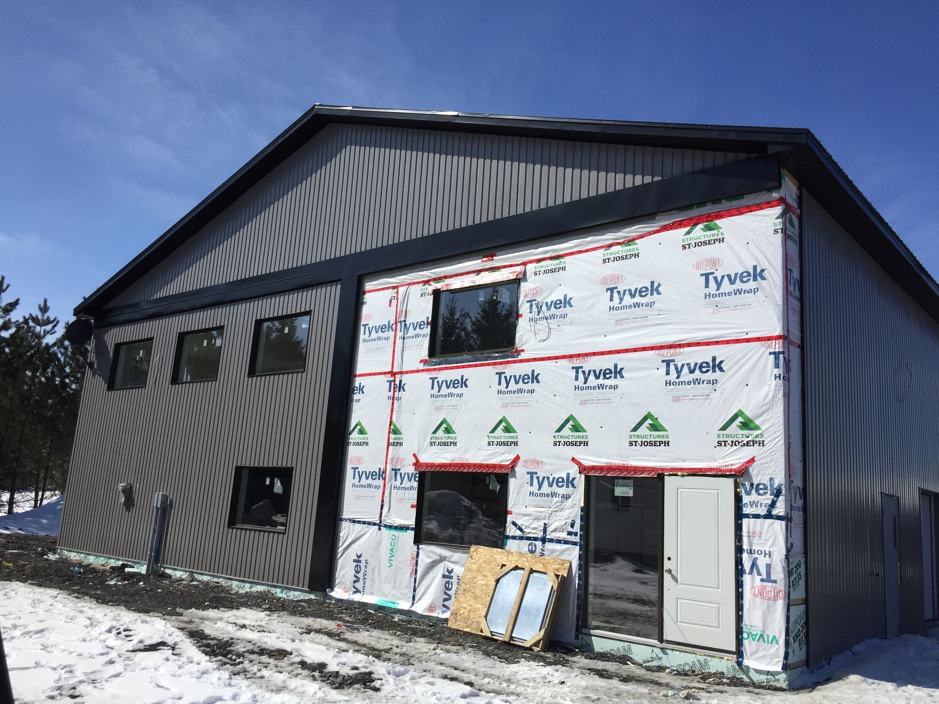 Bâtiment métallique gris en construction ; fenêtres, porte et pare-vapeur Tyvek visibles. Neige au sol.