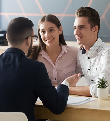 Man in suit with young couple at a table, discussing documents. All are smiling in an office setting.