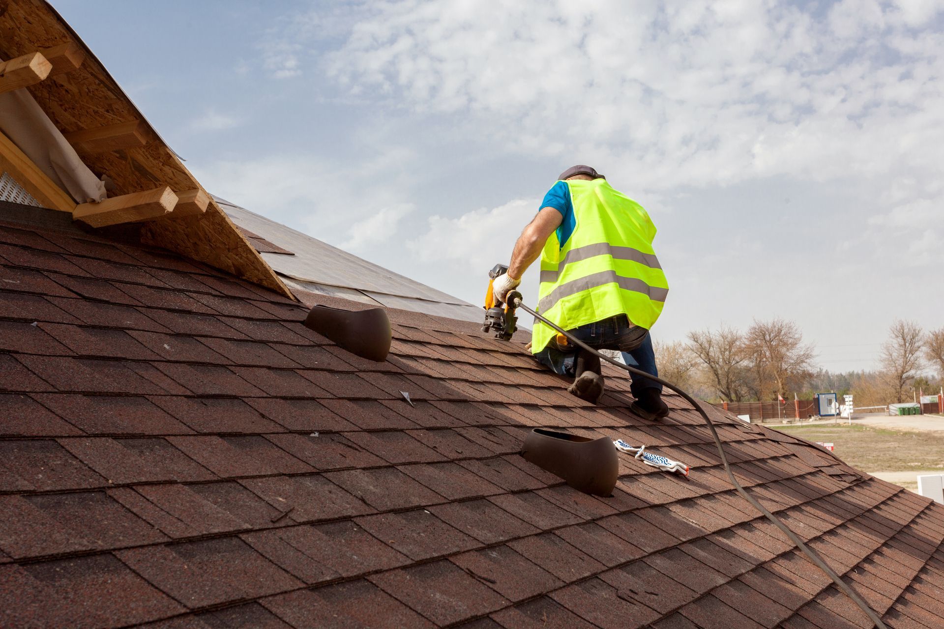 A roofer in a yellow vest secures shingles with a nail gun, safety ropes attached.