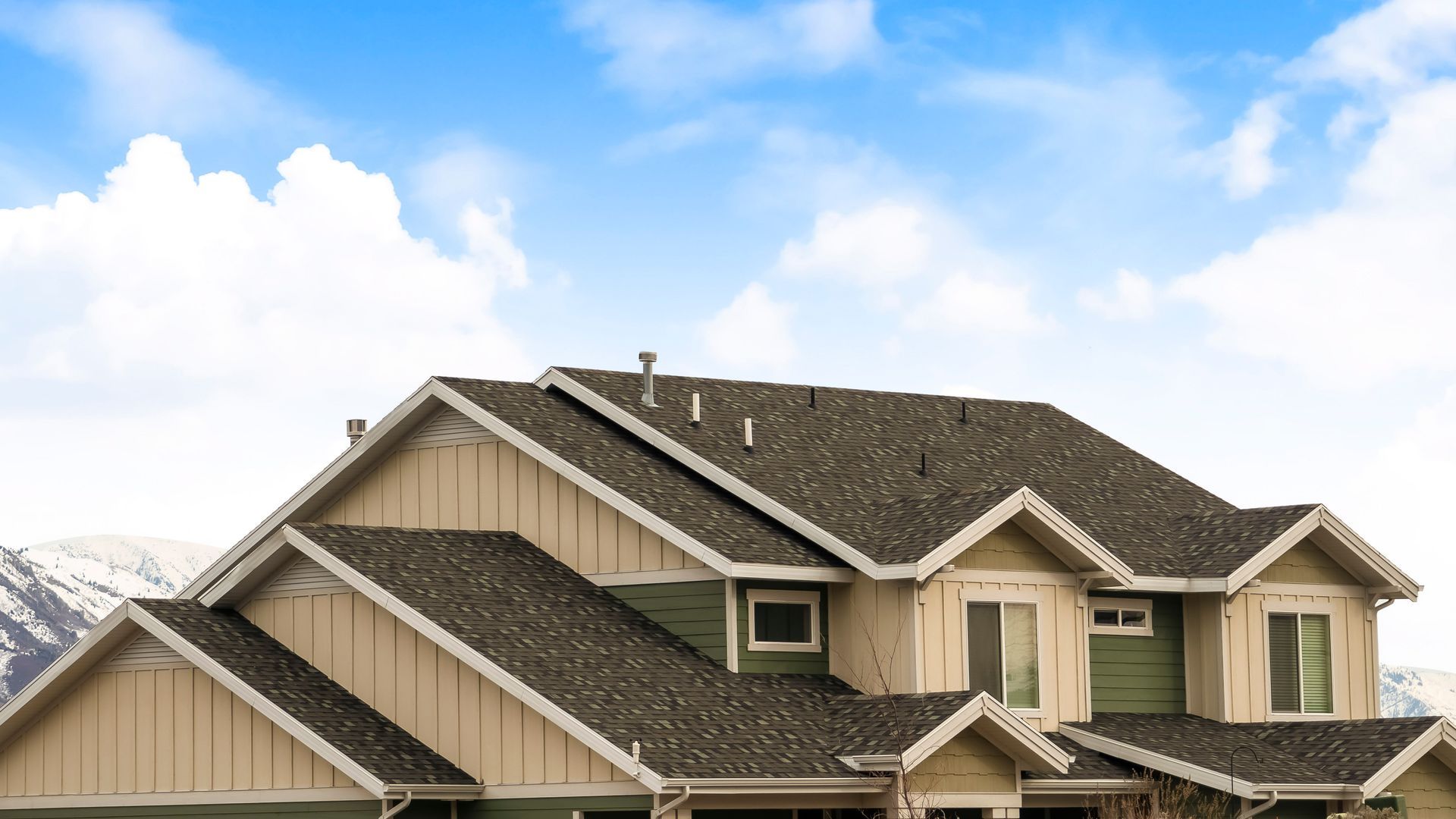 House with a dark gray roof, green siding, tan trim, and a blue sky with clouds in the background.
