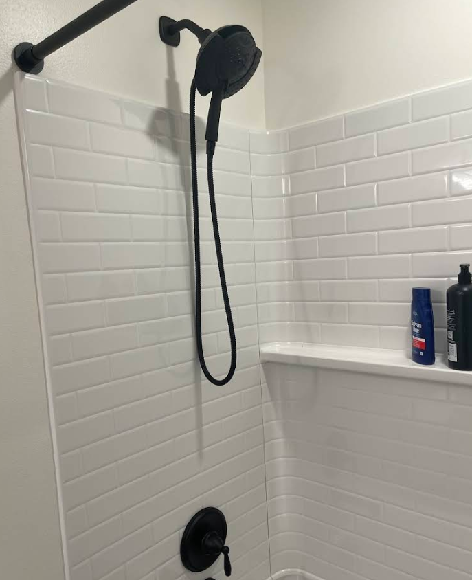 Black showerhead and fixtures in a white tiled shower, with shelf and shampoo bottles.