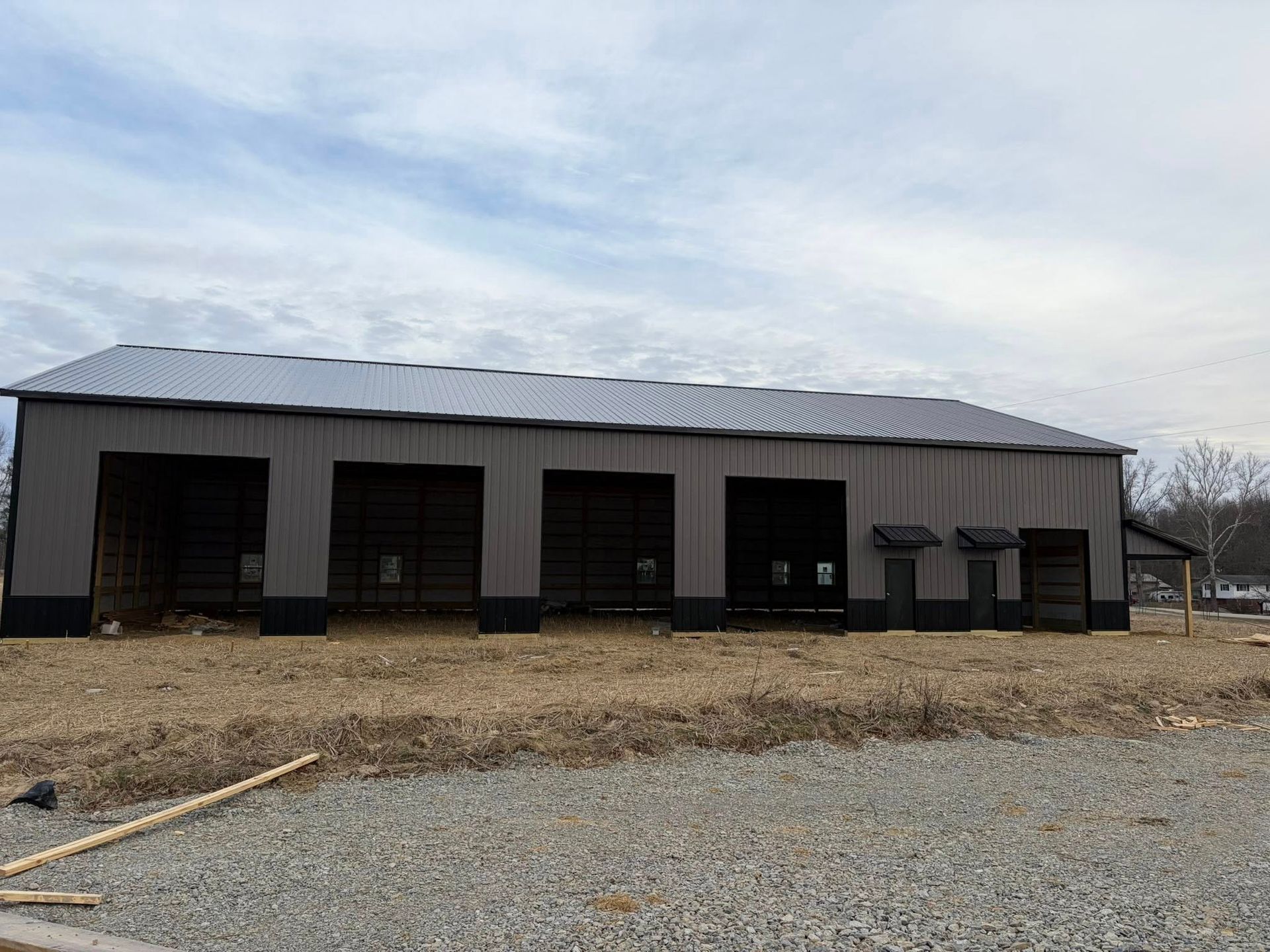Large gray metal barn with open bays on a gravel lot under a cloudy sky