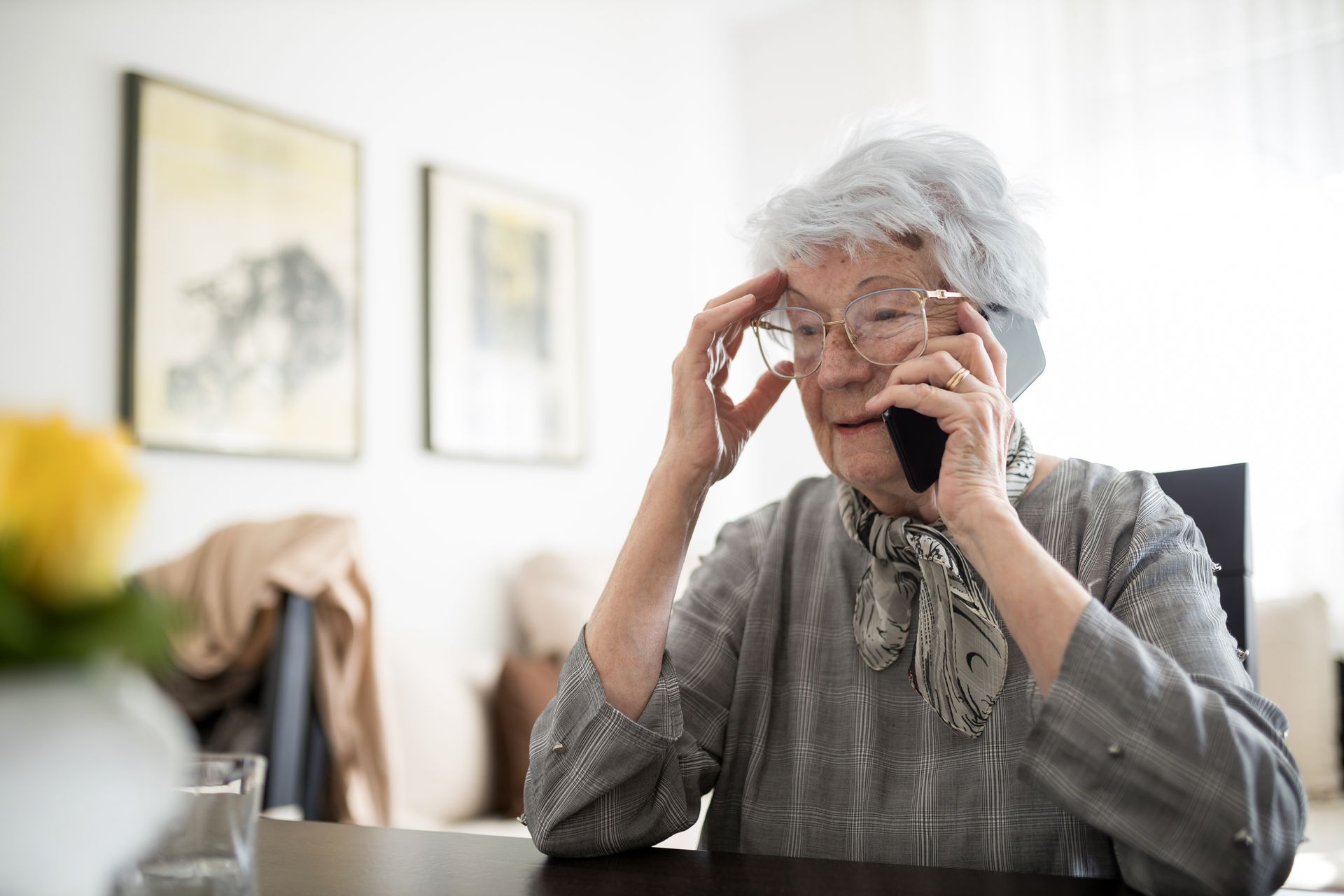 Elderly woman with glasses, on the phone, appears stressed, sitting indoors.