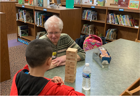 Child and elderly adult playing Jenga at a library table, with bookshelves and a water bottle nearby