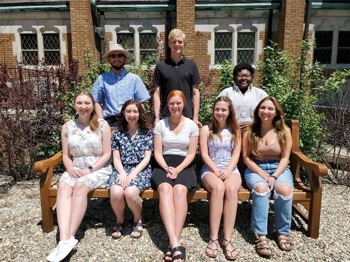 Eight people posing outdoors on benches in front of a brick building and garden.