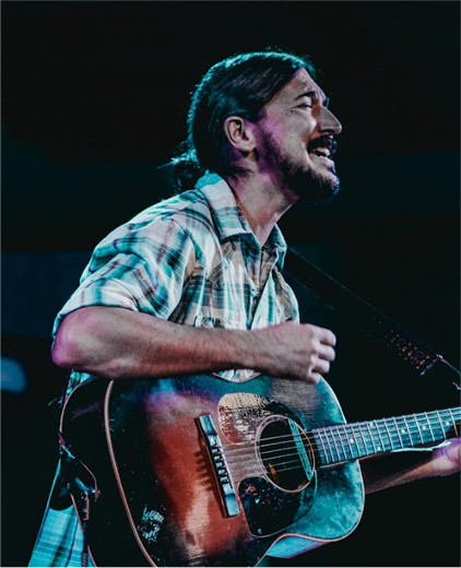 Singer playing acoustic guitar on stage, smiling under blue lighting