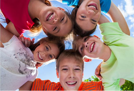 Five smiling kids leaning in a circle outdoors against a blue sky