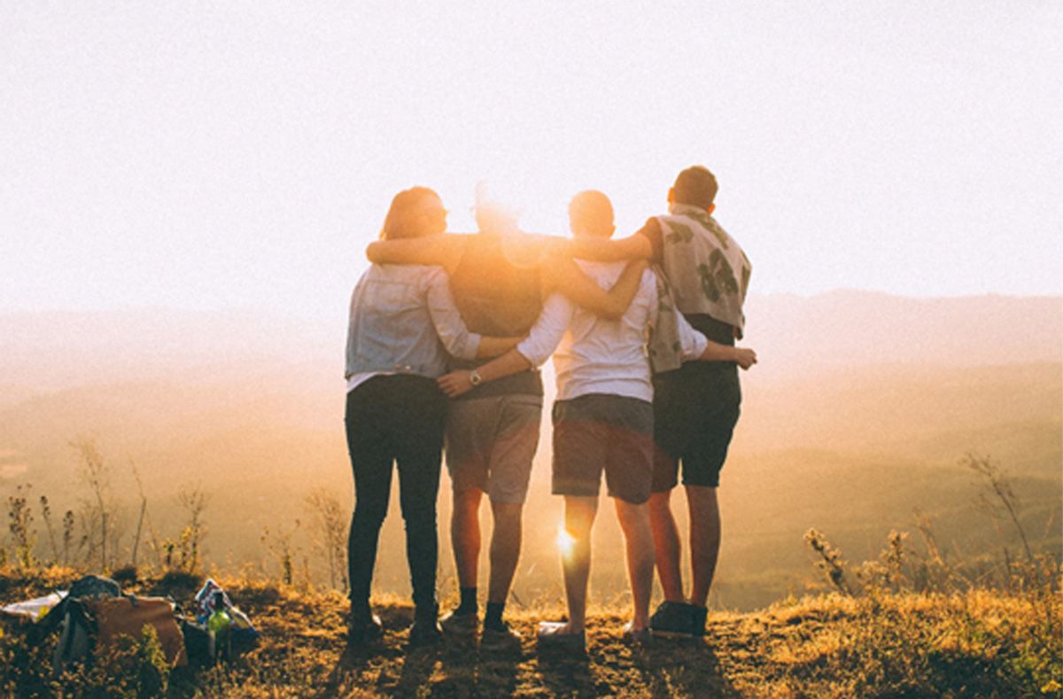 Four friends standing arm-in-arm in a sunlit field at sunrise, facing the horizon.