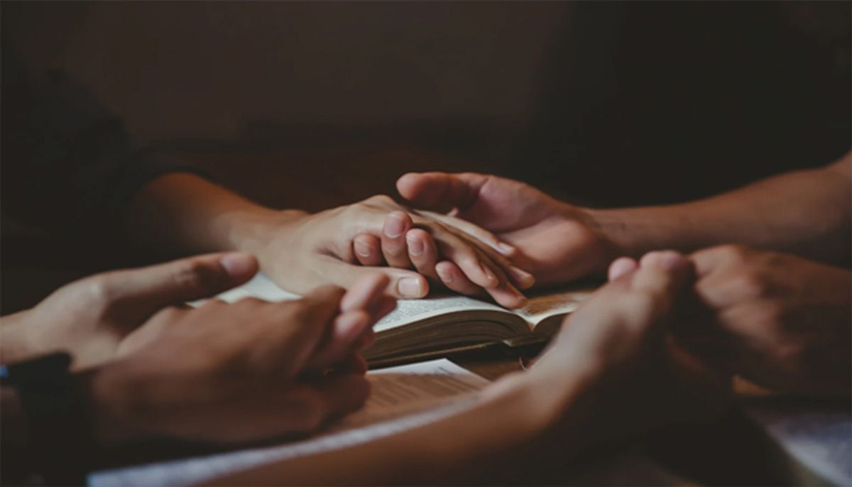 Hands gathered in a circle over a dimly lit table, suggesting a group meeting or prayer