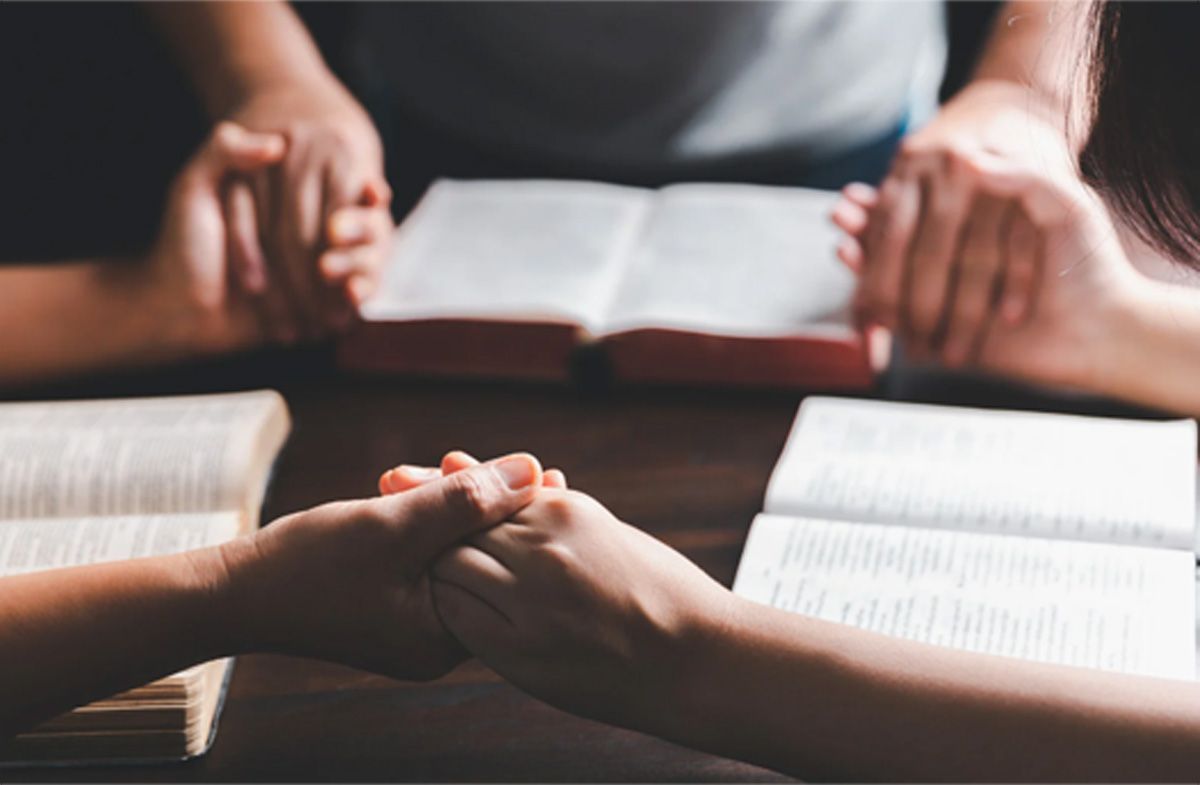 Two people holding hands across open books on a table, suggesting prayer or study