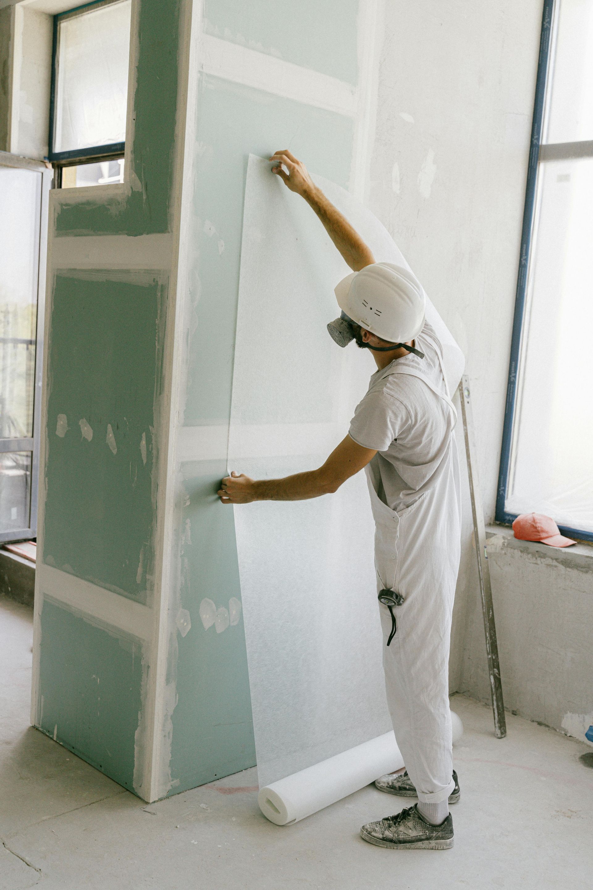 Man in white overalls and mask applies paper to a drywall column indoors.