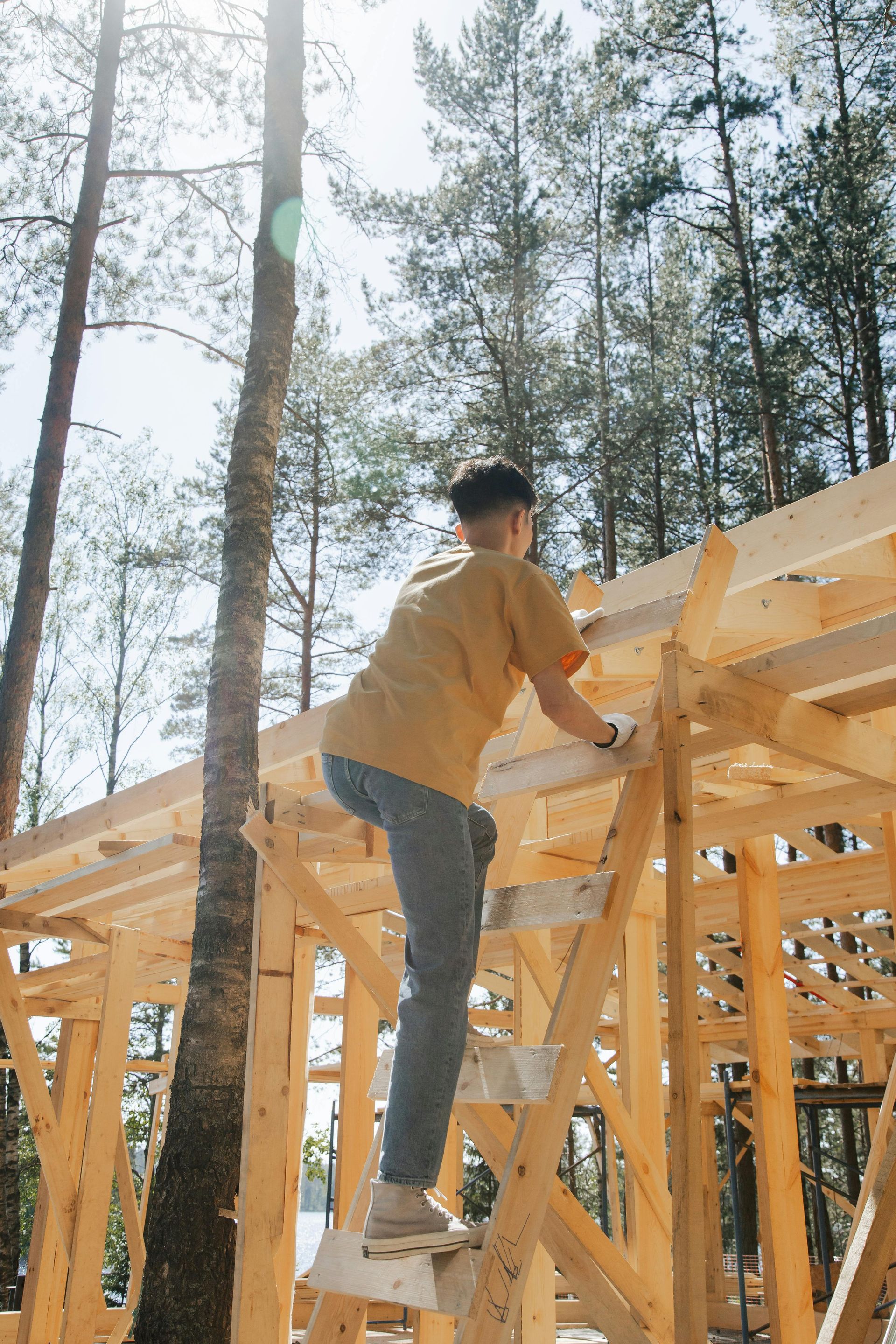 Man on a ladder, constructing a wooden building frame outdoors. He wears yellow shirt, gray pants.