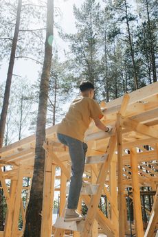Man on a ladder, constructing a wooden building frame outdoors. He wears yellow shirt, gray pants.