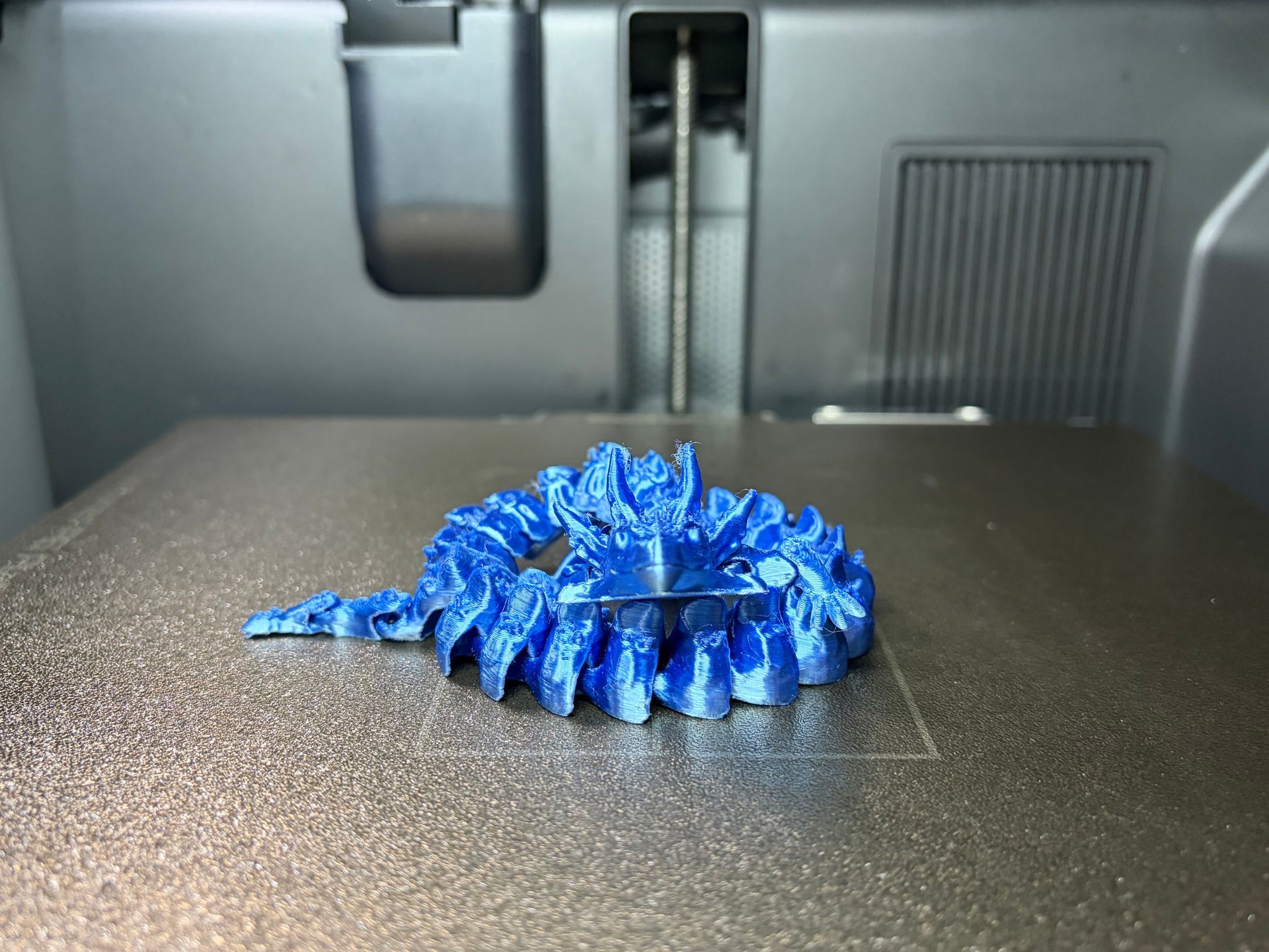 A partially printed blue 3D model with rough, unfinished textures sits on a 3D printer's heated print bed.