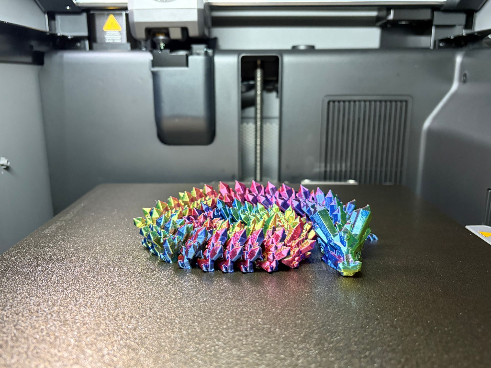 A colorful, rainbow-gradient 3D-printed articulated dragon toy resting on the textured print bed of a 3D printer.