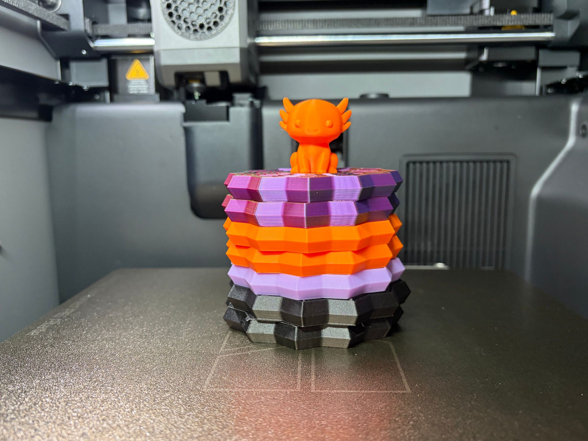 A stack of colorful, geometric 3D-printed blocks topped with a small orange axolotl figure inside a 3D printer.