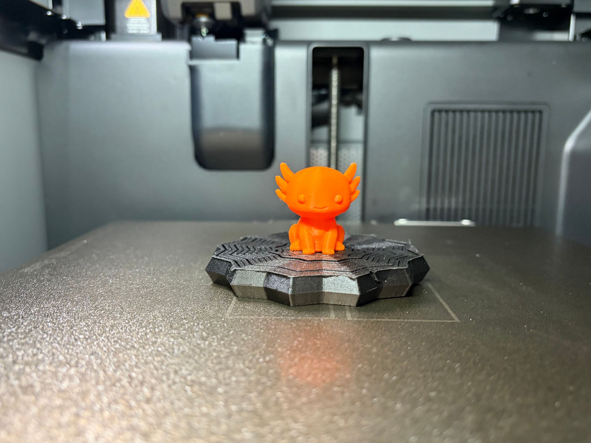 A small, orange 3D-printed axolotl figurine sits on a textured, dark gray circular platform inside a 3D printer.
