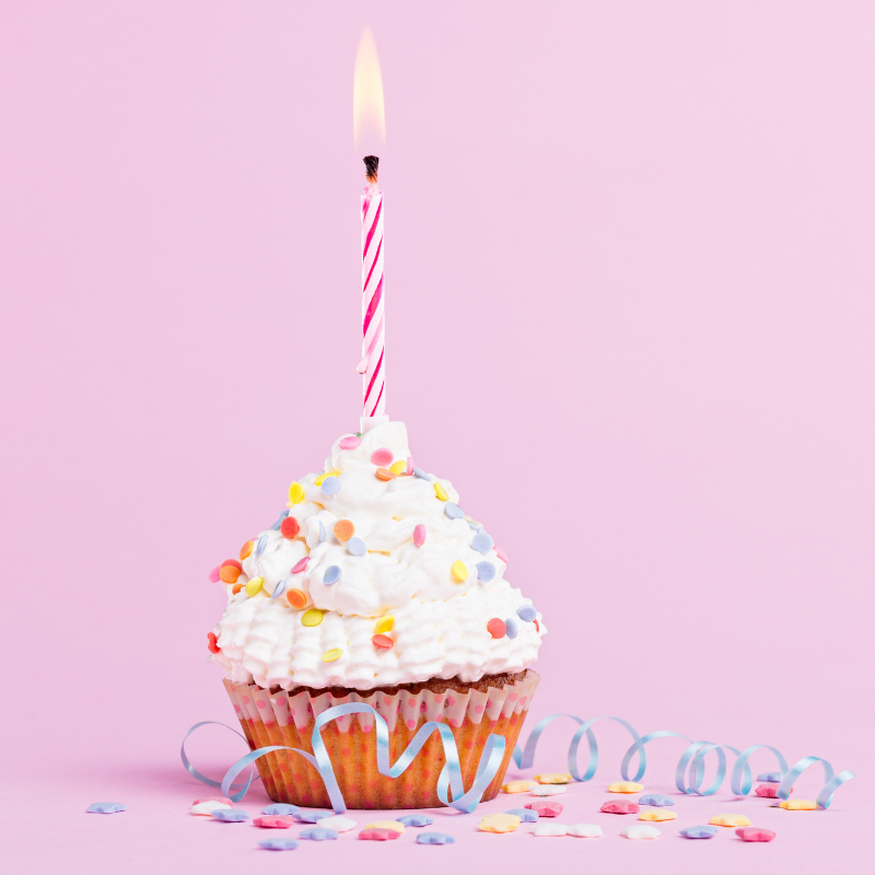 A birthday cupcake with a lit candle and sprinkles on a pink background.