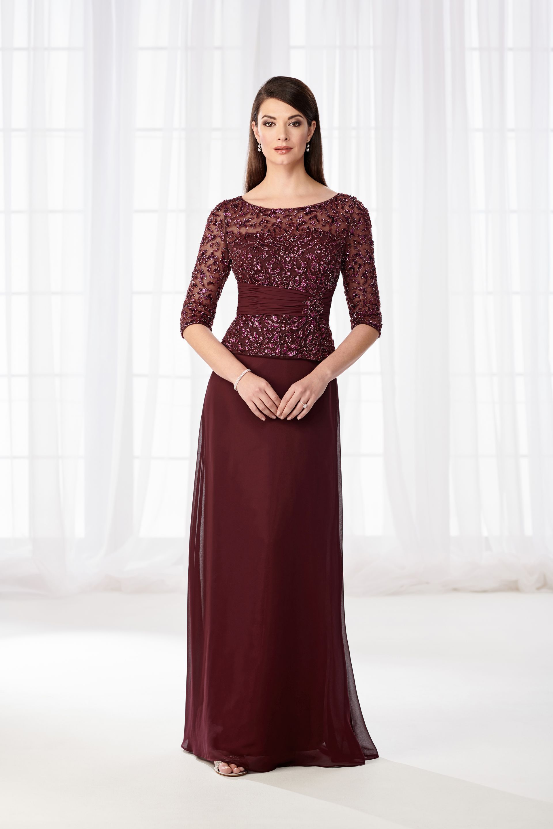 Woman in a burgundy formal gown with lace bodice, posing in front of sheer curtains.