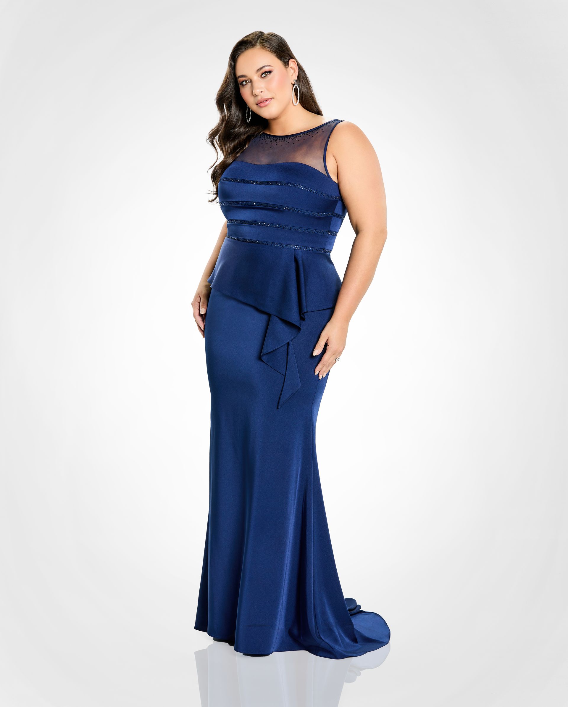 Woman in a navy blue formal gown, posed, looking at the viewer. Dress has a sheer neckline and a cascading ruffle.
