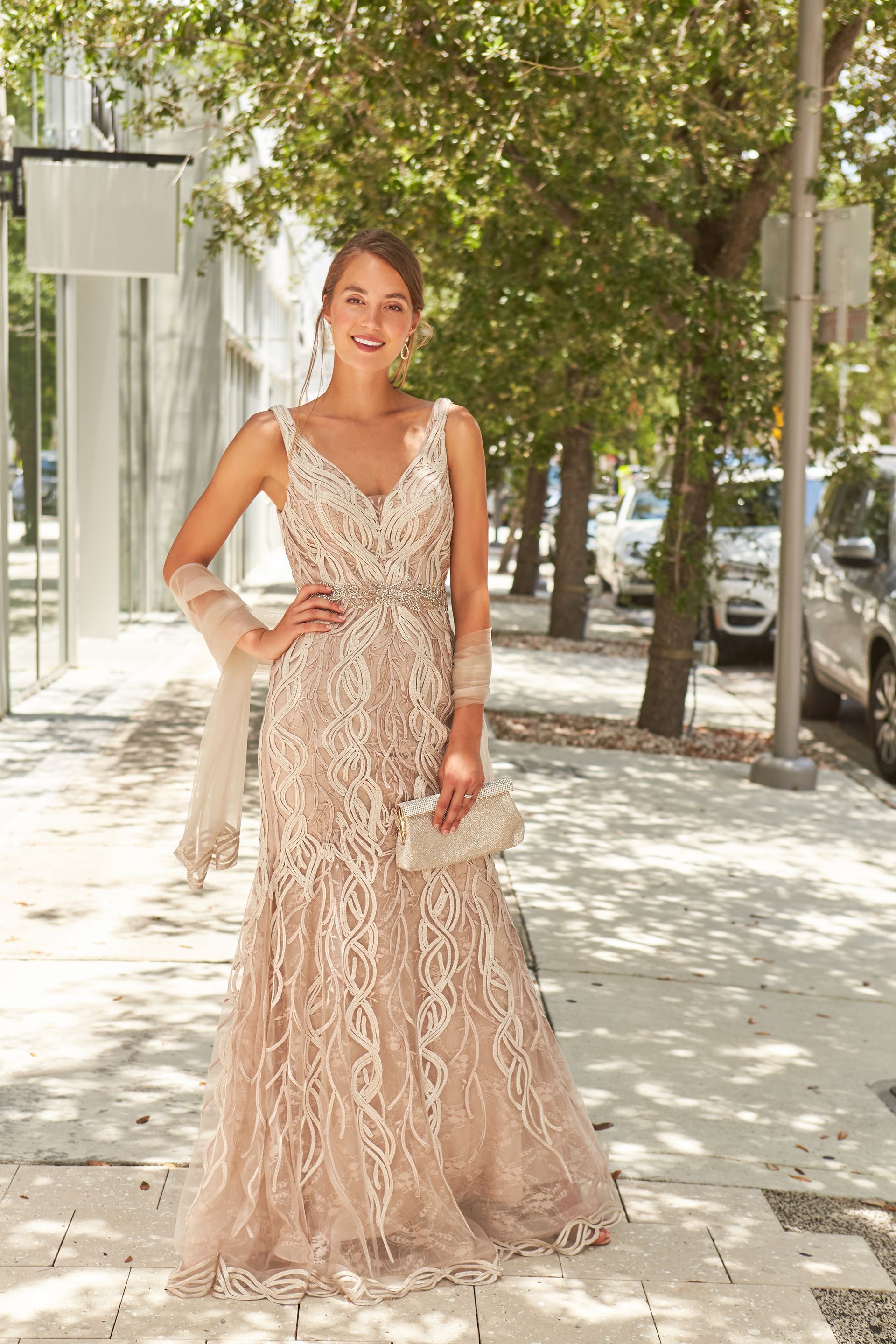 Woman in a gold lace gown with a shawl and clutch, standing outside on a sunny day.