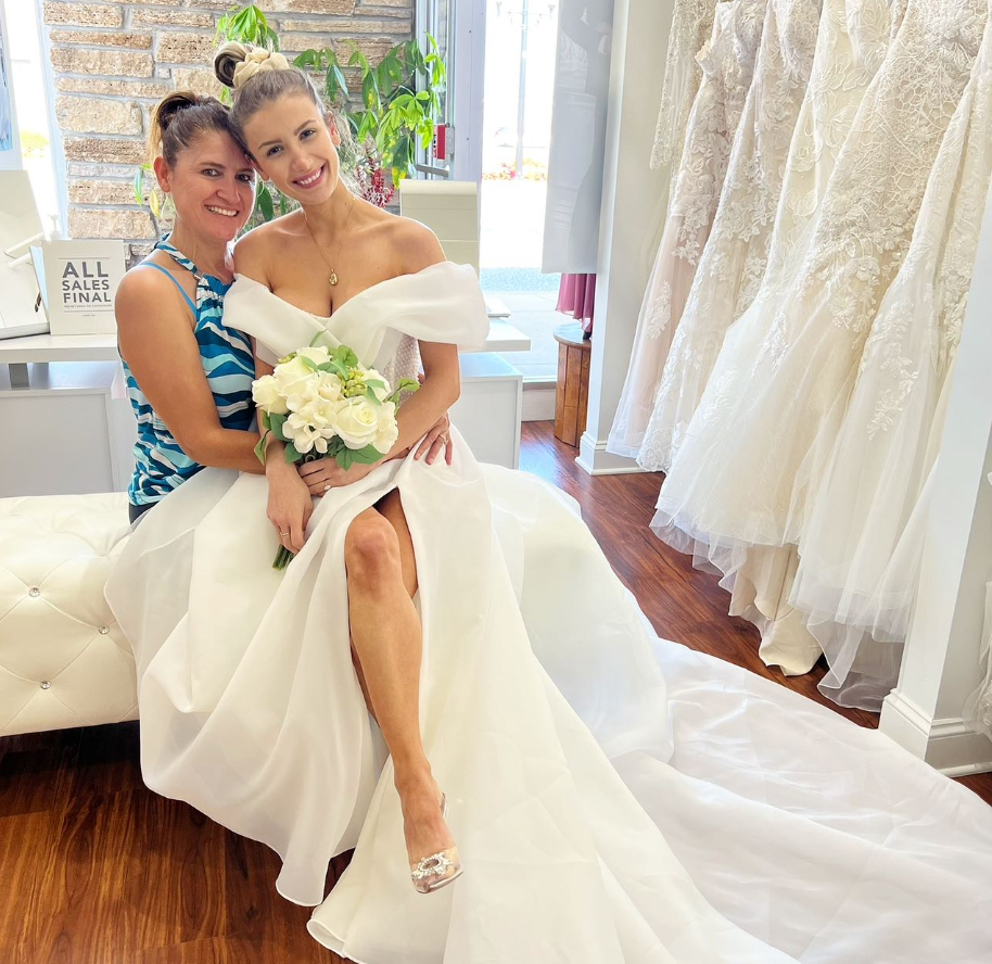 Woman in a white off-the-shoulder wedding dress smiles, holding flowers with a friend in a bridal shop.