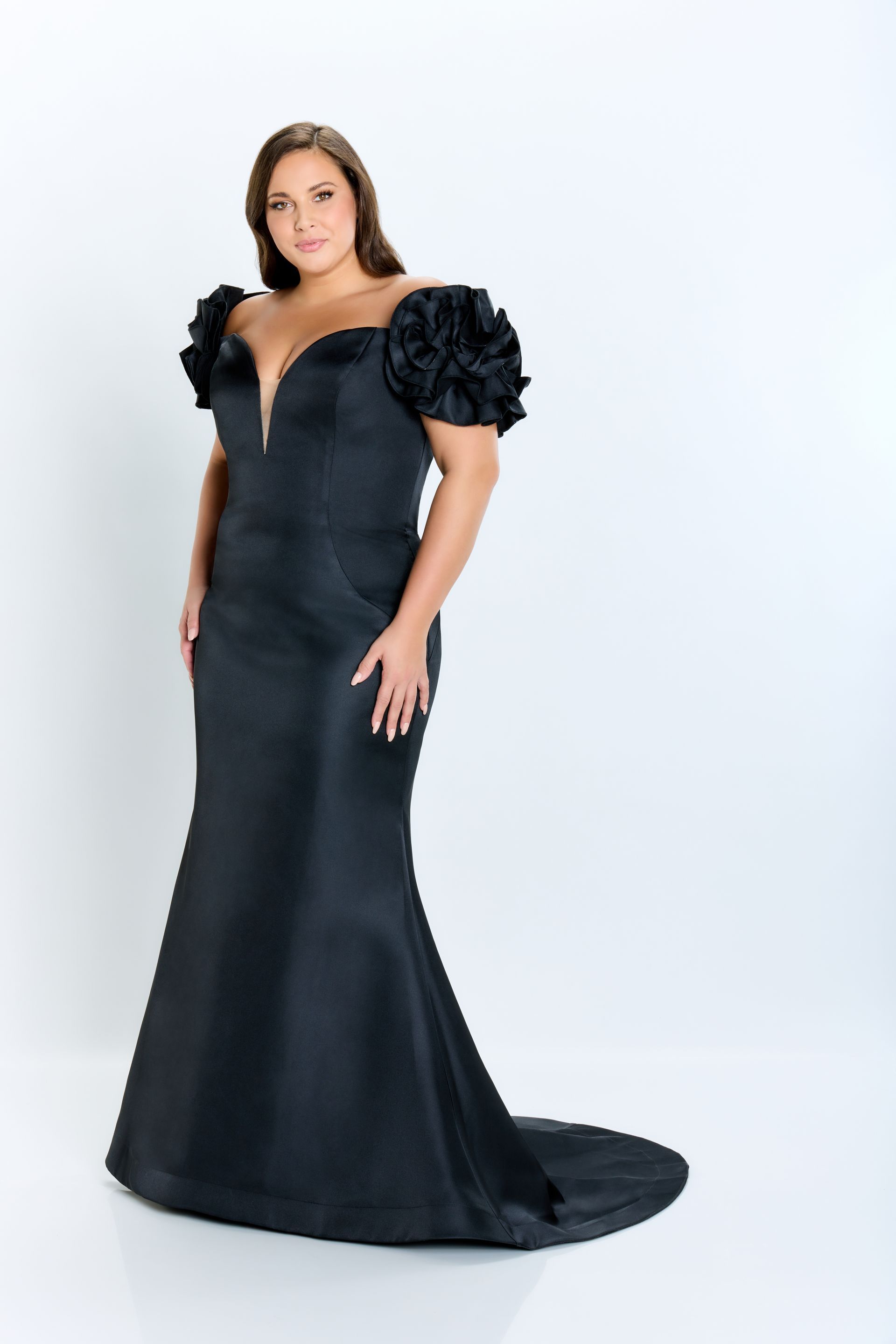 Woman in a black evening gown with floral shoulder details, posing against a white background.