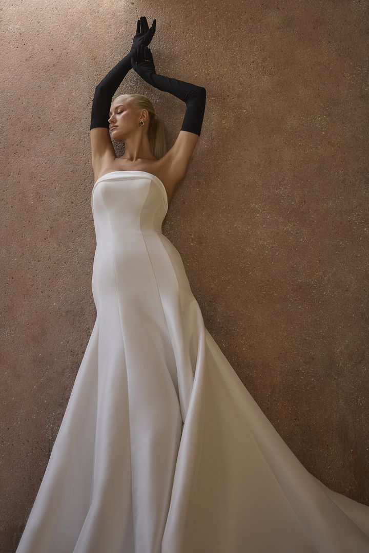 Woman in white strapless gown and black gloves, arms raised, posing against a textured brown wall.