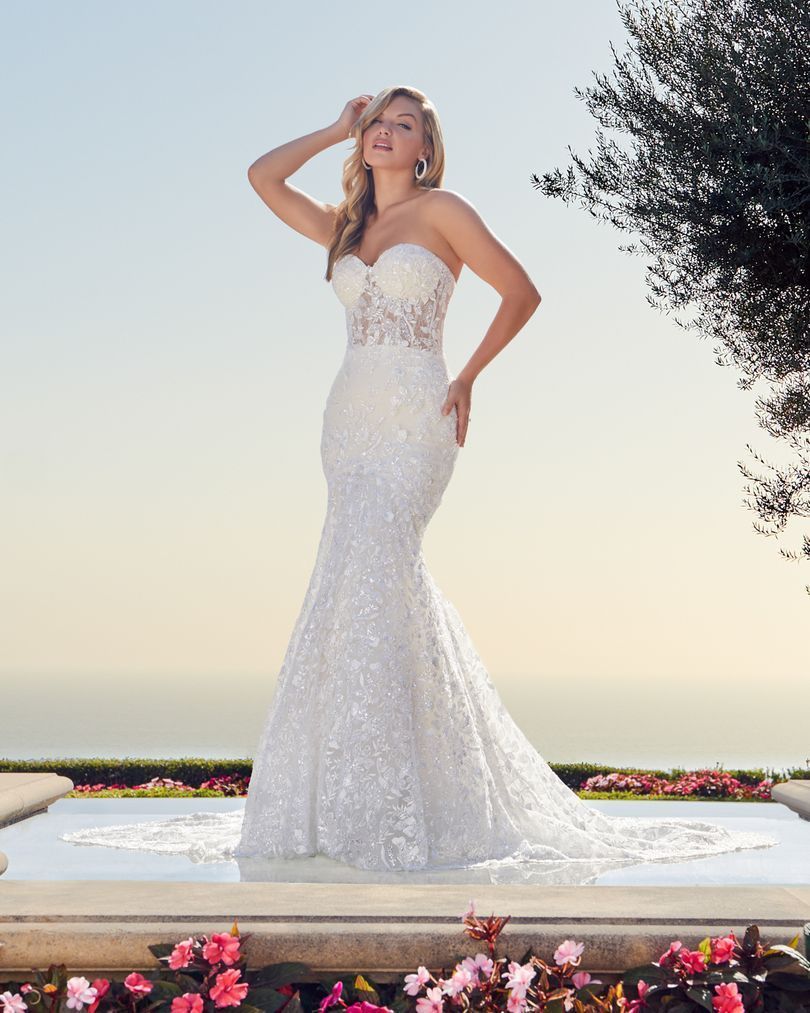 Woman in strapless white sequined wedding dress, posing outdoors near water, arms raised.