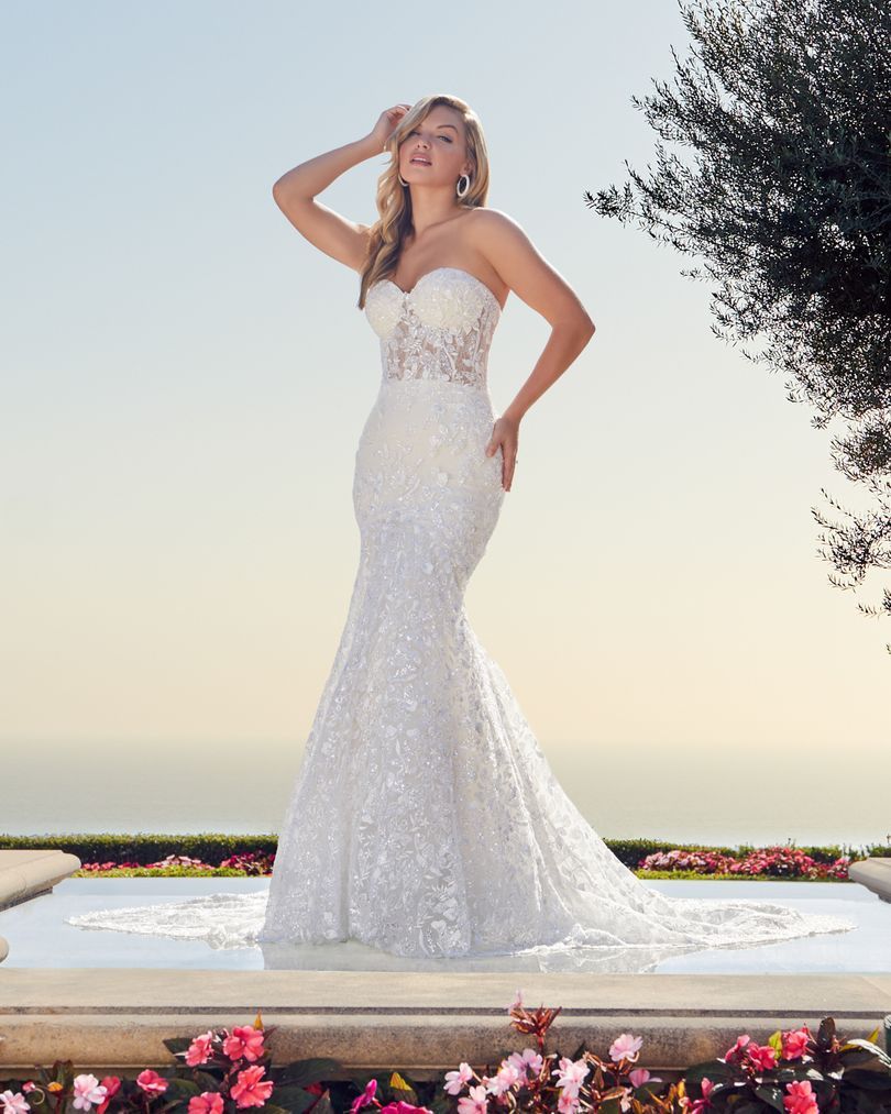 Blonde woman in strapless, sequined mermaid wedding dress, posing outdoors near water.