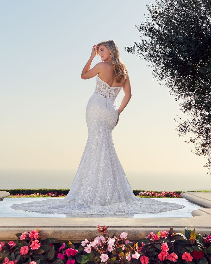 Blonde woman in strapless, sequined mermaid wedding dress, posing outdoors near water.