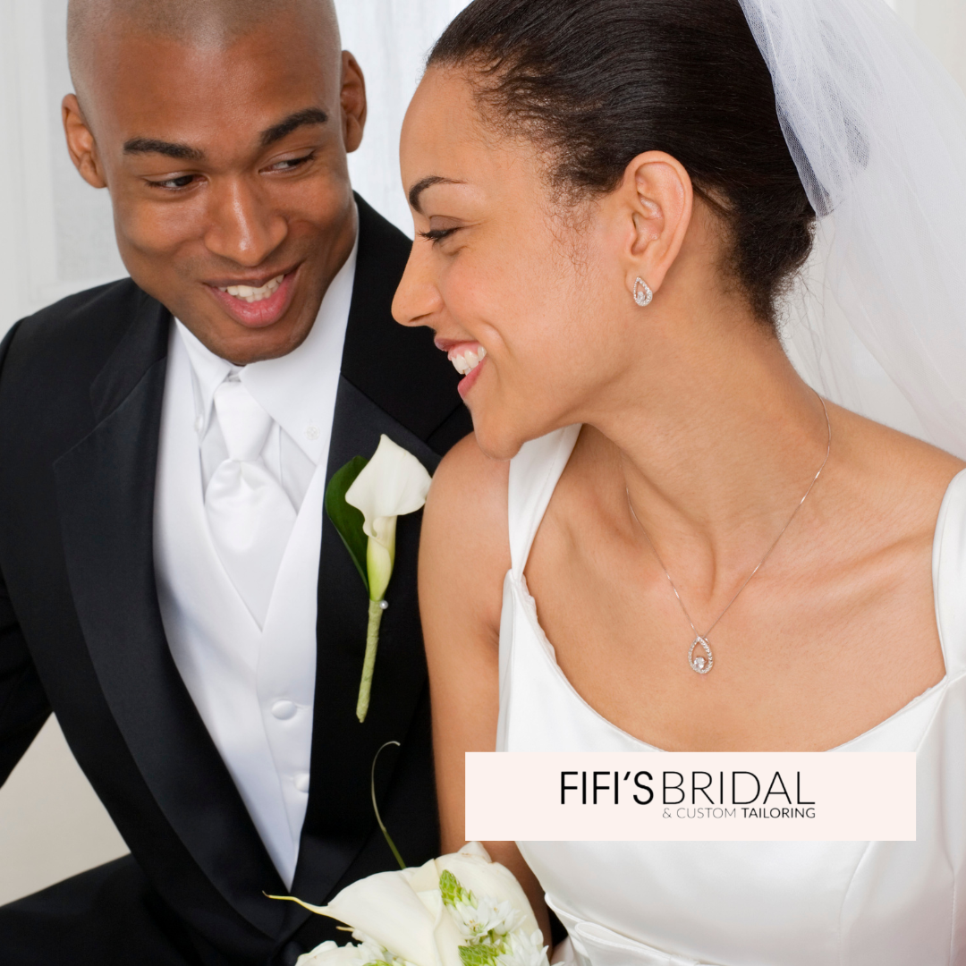 Bride and groom smiling at each other on their wedding day. She wears a white dress and veil, he wears a black suit.