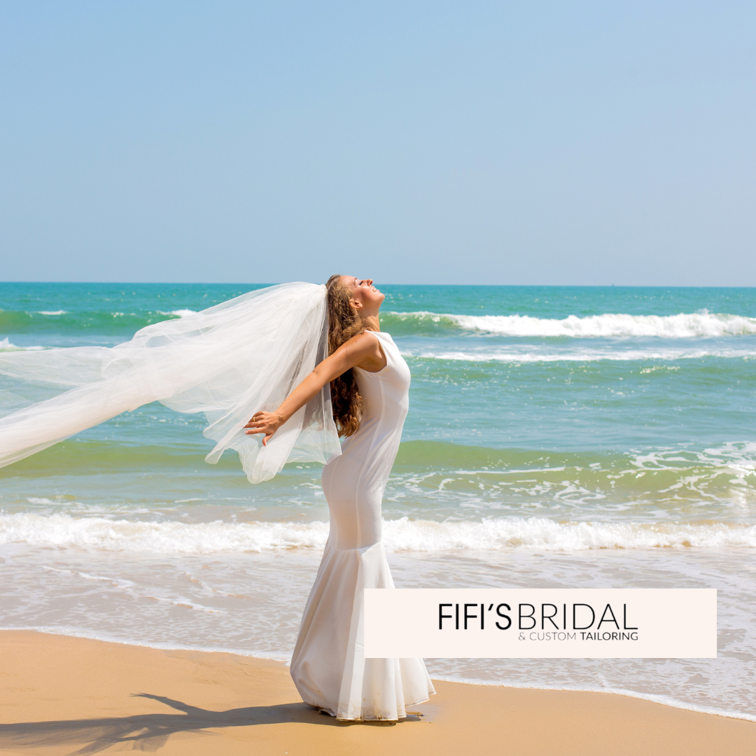 Bride on beach, arms outstretched, veil billowing, ocean background.