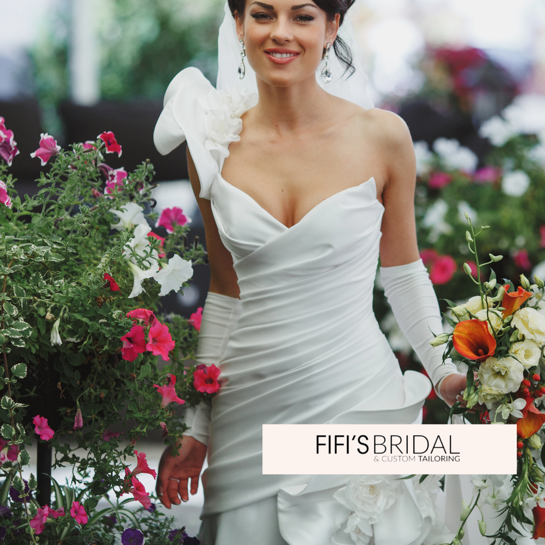 Bride in white strapless gown holds a bouquet, smiles. Surrounded by flowers, Fifi's Bridal logo present.