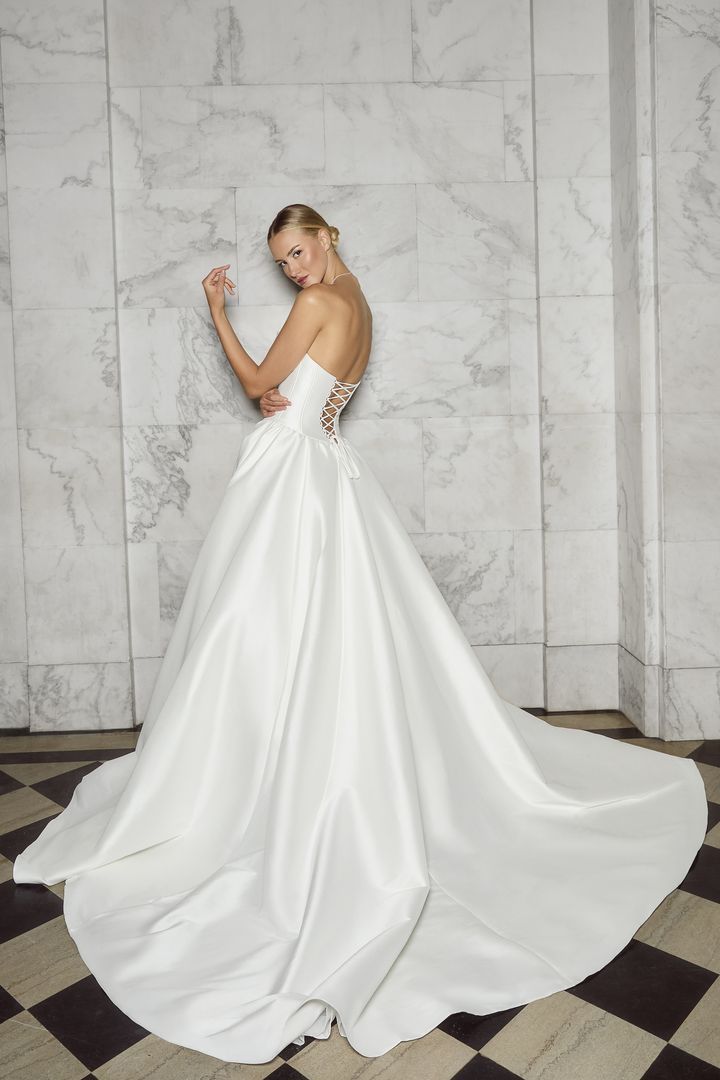 Woman in a white satin wedding gown, posing against a marble wall.