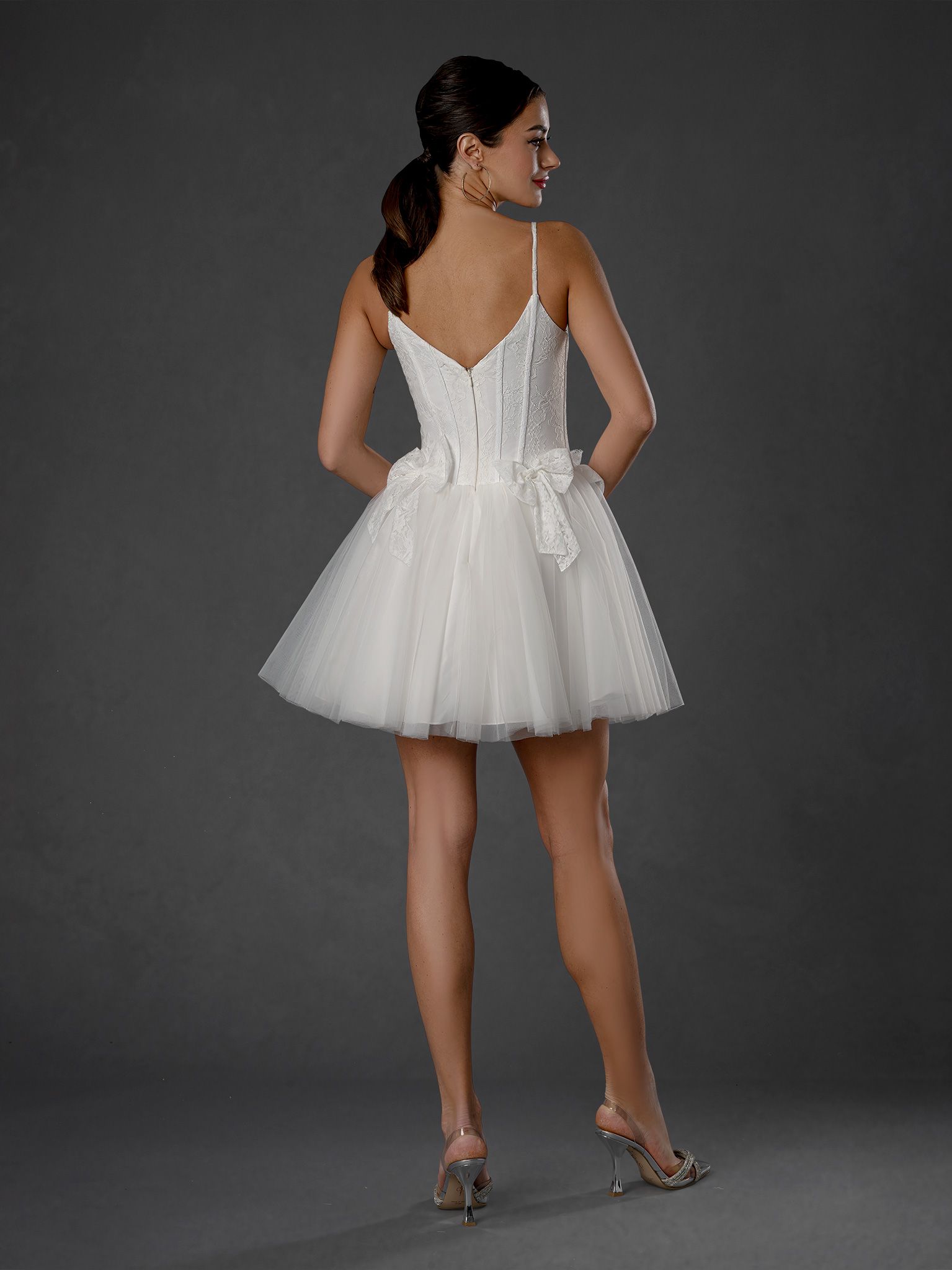 Woman in short white wedding dress, posing against gray backdrop.