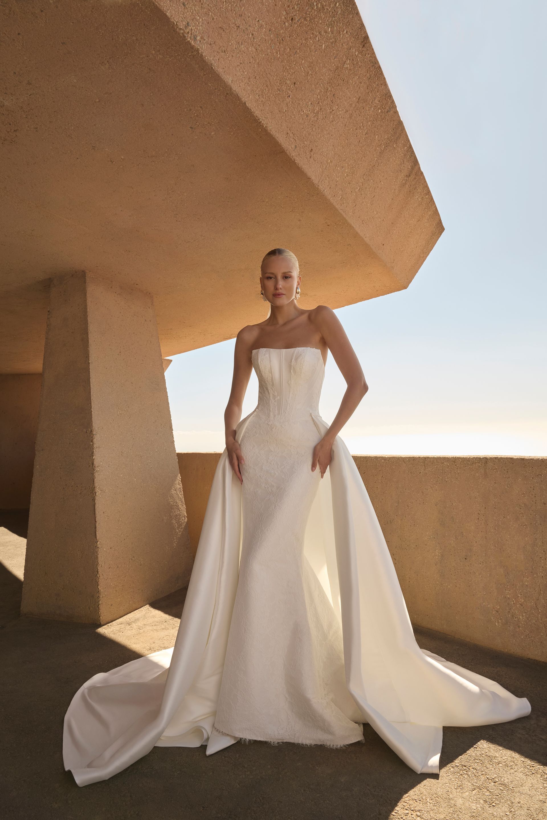 Woman in a strapless wedding dress with a long train, standing on a balcony with a concrete structure.