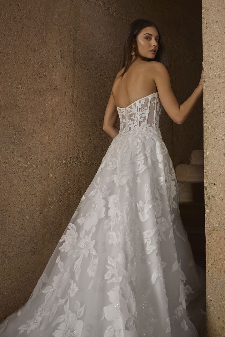 Woman in strapless white lace wedding dress, train on stone wall background.
