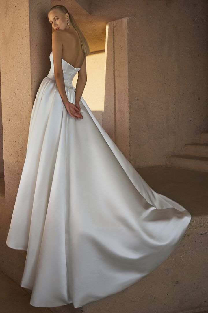 Woman in a white satin wedding gown, leaning against a stone wall, back view.