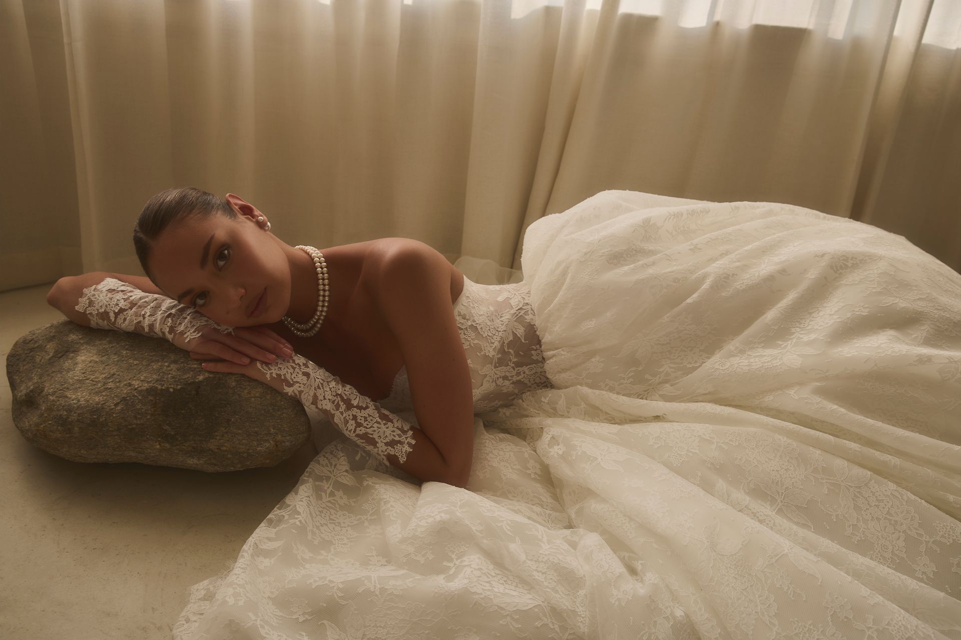 Woman in a white lace dress and gloves, lying on a stone, looking at the camera.