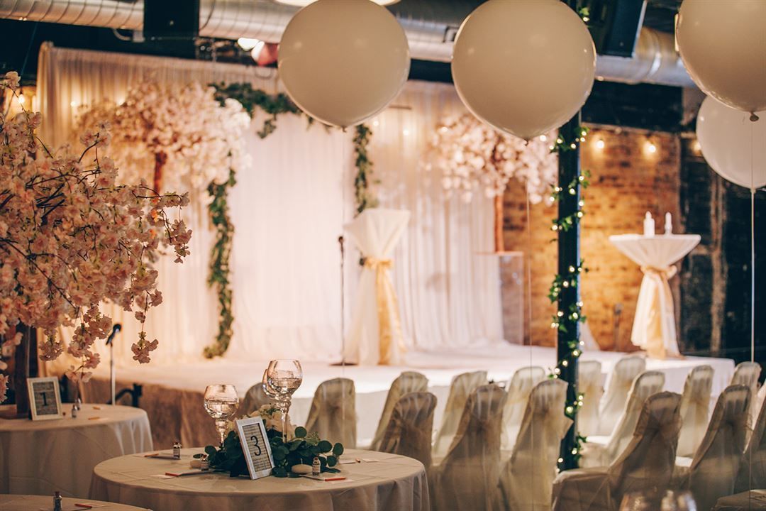 Wedding reception stage with white draped backdrop, floral decorations, balloons, and tables set for guests.