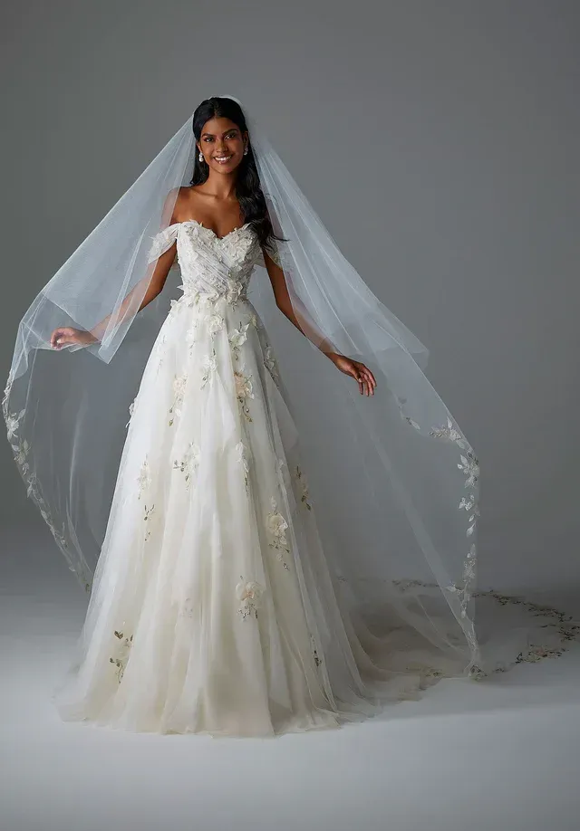A person smiles while wearing an off-the-shoulder, floral-appliqué bridal gown and a long veil against a gray background.