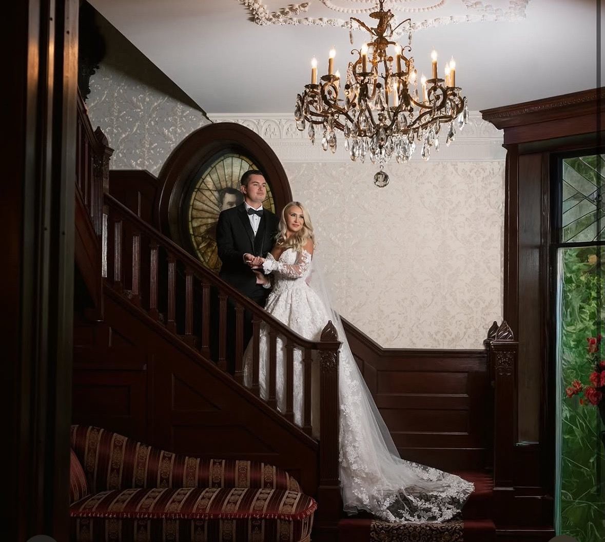 Couple on a grand staircase, bride in long white gown, groom in tuxedo. Elegant room, ornate chandelier.