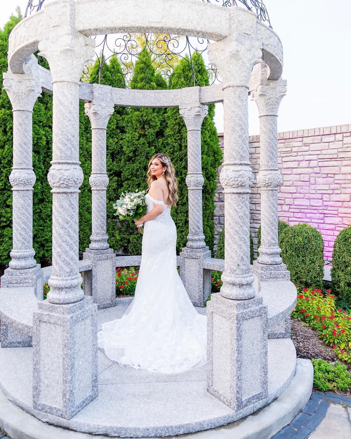 Bride in white wedding dress holding bouquet, posing in a stone gazebo.