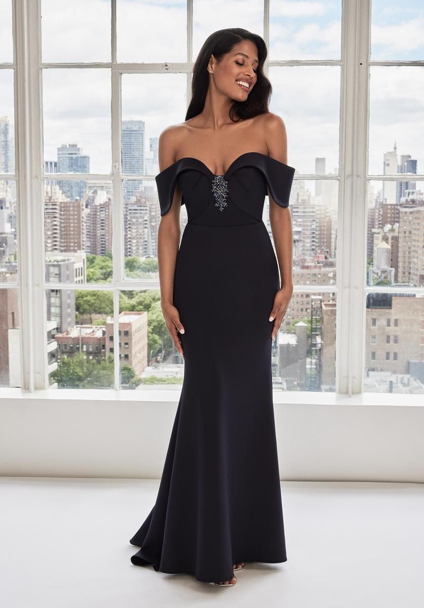 Woman in a black off-the-shoulder gown, smiling, near a window with city view.